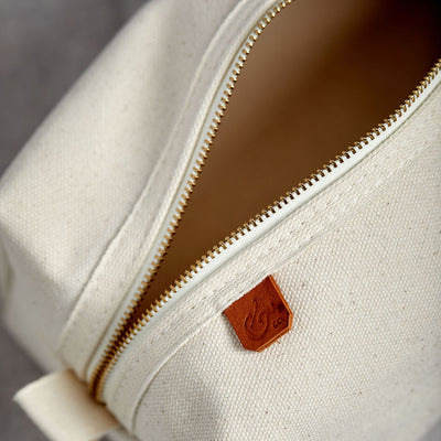 Overhead close-up of Dopp kit made from sturdy off-white canvas with metal zipper and leather detail. Classic small travel and toiletry bag.
