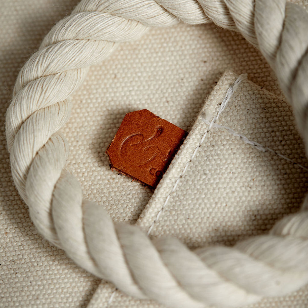 Close-up detail of minimalist, maritime-inspired white canvas tote bag. Thick cotton rope and handcrafted leather detail with stamped logo.